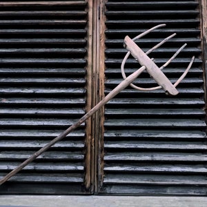 Puede incluir: Un tenedor de heno de madera con cuatro dientes, apoyado contra un conjunto de contraventanas de madera. El tenedor está desgastado y tiene un acabado natural.
