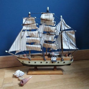 Puede incluir: Un modelo de barco de madera con velas blancas y aparejos negros. El barco está sobre una base de madera y tiene un casco verde claro.