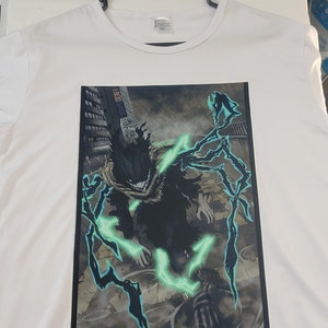 K&ouml;nnte beinhalten: Wei&szlig;es T-Shirt mit einem schwarzen und gr&uuml;nen Grafikdruck eines Charakters mit leuchtend gr&uuml;ner Energie. Der Charakter steht in einer Stadtkulisse mit hohen Geb&auml;uden im Hintergrund.