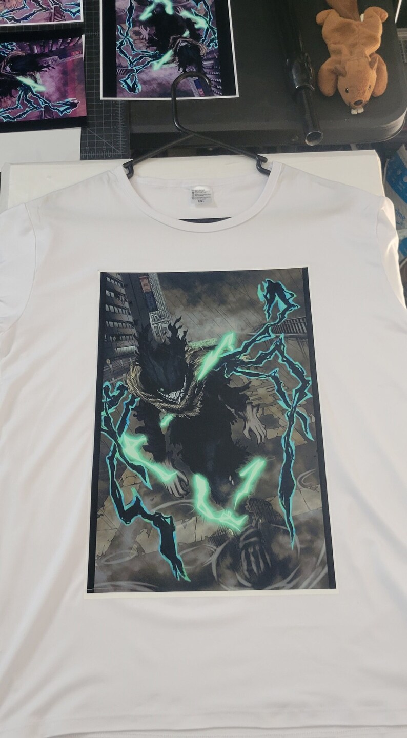 K&ouml;nnte beinhalten: Wei&szlig;es T-Shirt mit einem schwarzen und gr&uuml;nen Grafikdruck eines Charakters mit leuchtend gr&uuml;ner Energie. Der Charakter steht in einer Stadtkulisse mit hohen Geb&auml;uden im Hintergrund.