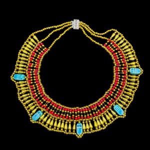 May include: A gold-coloured necklace with red and black accents, featuring turquoise-coloured scarab beetle-shaped pendants. The necklace has a layered design with small, round beads and a decorative clasp.