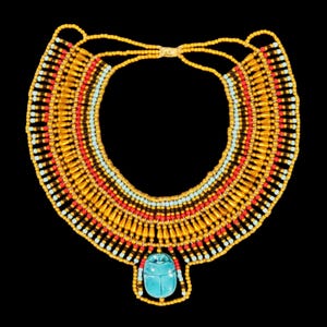 Op de afbeelding: Een goudkleurige kralen ketting met rode, zwarte en lichtblauwe accenten. De ketting heeft een turquoise scarabee hanger. Het ontwerp doet denken aan oude Egyptische sieraden.