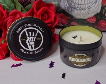 May include: A black candle tin with a white label that reads "Blood Red Plum". The candle is surrounded by purple glitter bats and a white ghost figurine. The candle tin lid has a black and white graphic of a skeleton hand with the text "Sweet Metal Skeleton Est. 2021 Goth & Art Designs."