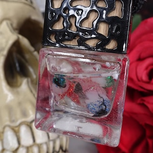 May include: A clear, square-shaped air freshener with a black top and a black cord. The air freshener contains colorful glitter and decorative elements. The background includes a skull and a red rose.