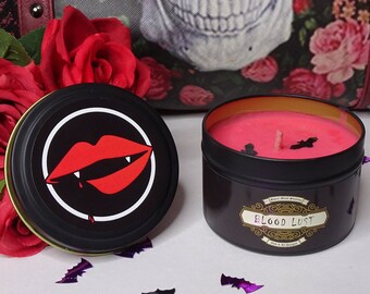 May include: A black candle tin with a pink candle inside. The candle has a black bat and a single wick. The label on the candle says "Blood Lust". The tin has a black and red image of a vampire's mouth with fangs on the lid.