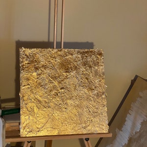 May include: A gold-leaf painting on a wooden easel. The painting has a textured surface and is a rich, golden color.
