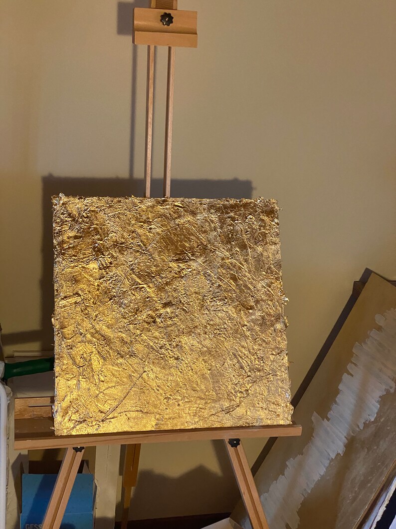 May include: A gold-leaf painting on a wooden easel. The painting has a textured surface and is a rich, golden color.