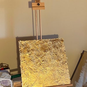 May include: A gold-toned canvas painting on an easel. The canvas is textured and has a rough, uneven surface. The painting is in a contemporary style.