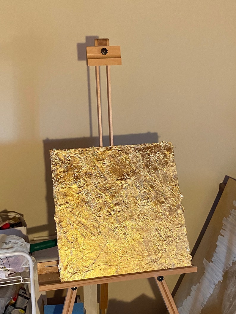 May include: A gold-toned canvas painting on an easel. The canvas is textured and has a rough, uneven surface. The painting is in a contemporary style.