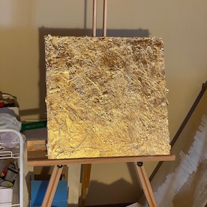 May include: A gold-leaf painting on a wooden easel. The painting has a textured surface and is set against a beige wall.