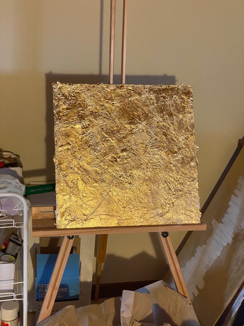 May include: A gold-leaf painting on a wooden easel. The painting has a textured surface and is set against a beige wall.