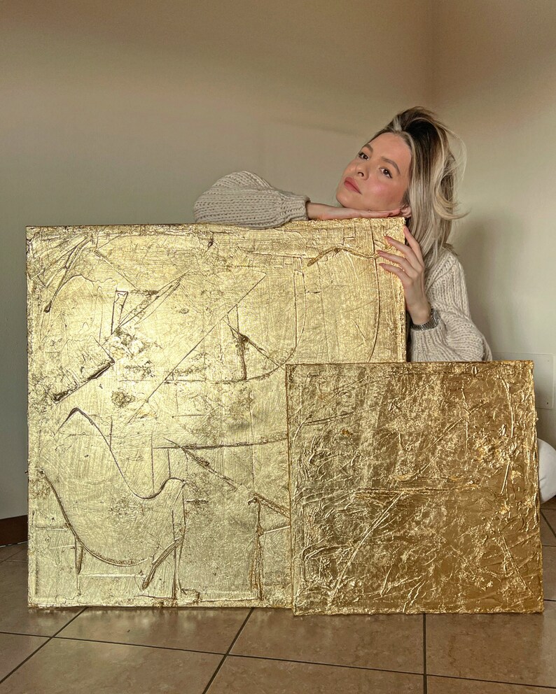 May include: Two gold leaf abstract paintings with textured surfaces. The larger painting has a more abstract design, while the smaller painting has a more geometric design.