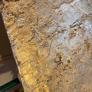 May include: A textured gold canvas painting with a white abstract design. The canvas is mounted on a wooden easel.