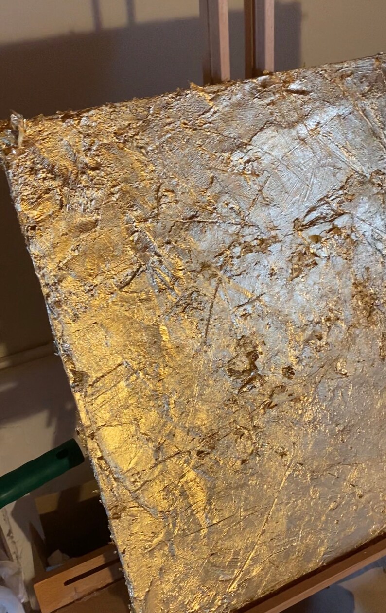May include: A textured gold canvas painting with a white abstract design. The canvas is mounted on a wooden easel.