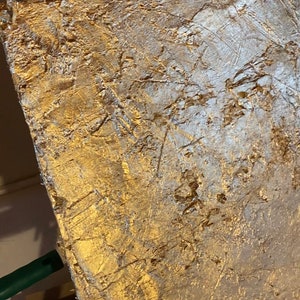 May include: A gold and silver abstract painting on canvas with a textured surface. The painting is on an easel.