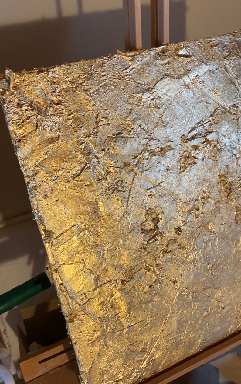 May include: A gold and silver abstract painting on canvas with a textured surface. The painting is on an easel.
