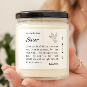 May include: A white candle with a black lid and a label that says "Secret Gardens Design" and "Sarah". The label also includes a dove with an olive branch in its beak and the text "Don't you be afraid, for I am with you. Don't be dismayed, for I am your God. I will strengthen you. Yes, I will help you. Yes, I will uphold you with the right hand of my righteousness. Isaiah 41:10".