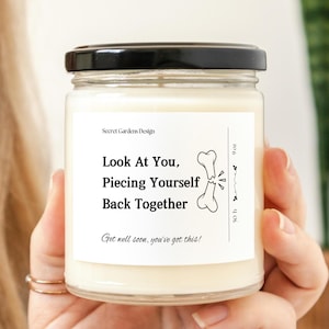 May include: A white candle with a black lid and a label that reads "Look At You, Piecing Yourself Back Together" with a broken bone graphic. The text below the graphic reads "Get well soon, you've got this!"