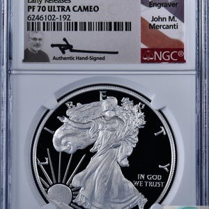 May include: A 2021 W Eagle S$1 silver coin, graded PF 70 ULTRA CAMEO, in a protective case. The coin shows Lady Liberty and the inscription "IN GOD WE TRUST". The holder displays details about the coin and the engraver, John M. Mercanti. The coin is hand-signed.