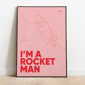 May include: Framed pink poster with a red rocket illustration. The words "I'M A ROCKET MAN" are in red, with the year "1972" at the top. Additional text is also present, all set against a pink background.