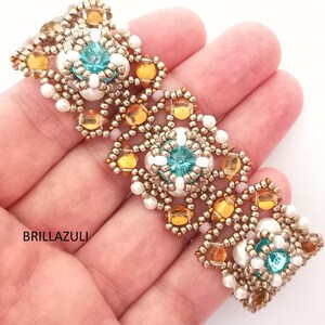 May include: A beaded bracelet with gold, white, and turquoise beads. The bracelet is made with a repeating pattern of small beads and larger beads. The bracelet is shown on a hand.