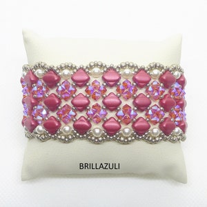 Könnte beinhalten: Ein rotes und weißes Perlenarmband mit geometrischem Muster. Das Armband liegt auf einem weißen Kissen mit dem aufgedruckten Wort "BRILLAZULI".