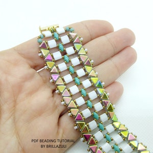 May include: A multi-colored beaded bracelet with white, turquoise, pink, and gold beads. The bracelet is made with small, square and triangular beads. The bracelet is on a hand, and the text "PDF BEADING TUTORIAL BY BRILLAZULI" is visible below the bracelet.