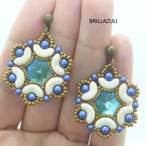 Può includere: Un paio di orecchini pendenti con un design a stella blu e bianco. Gli orecchini sono realizzati con piccole perline e un cristallo blu più grande al centro. Gli orecchini sono appesi a ganci in bronzo.