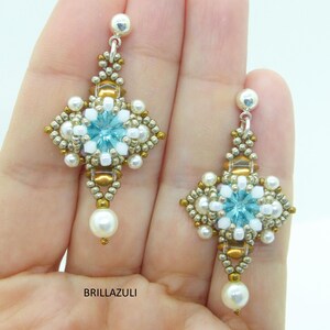 Puede incluir: Un par de pendientes colgantes con un diseño de cuentas azules y blancas. Los pendientes presentan un pequeño cristal azul en el centro de cada diseño. Los pendientes están suspendidos de ganchos plateados.