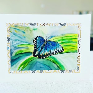 May include: A watercolor painting of a blue butterfly with black markings on its wings. The butterfly is perched on a green leaf with a light blue background.