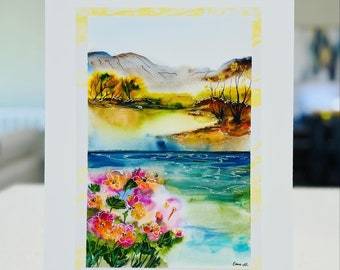 May include: Watercolor painting of a landscape with mountains, trees, a river, and flowers. The painting is framed in a white mat with a yellow border. The artist's signature is in the bottom right corner.