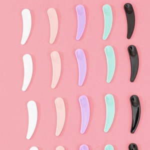 Può includere: Una collezione di spatole cosmetiche in vari colori, tra cui bianco, pesca, lavanda, verde menta e nero, disposte su uno sfondo rosa. Le spatole hanno un design curvo e allungato, ideali per l'applicazione di prodotti per la cura della pelle.
