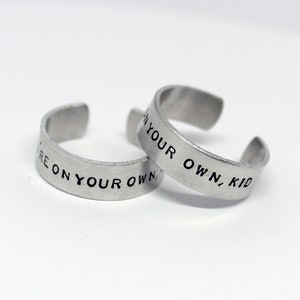 Può includere: Due anelli regolabili color argento. Ogni anello ha la frase "YOU'RE ON YOUR OWN, KID" stampata in lettere nere. Gli anelli sono su uno sfondo bianco.
