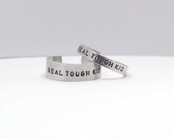Puede incluir: Dos anillos ajustables de color plateado con la frase "REAL TOUGH KID" estampada. Los anillos son de metal brillante y se muestran sobre una superficie blanca. El texto está en una fuente sencilla, sin serifa.