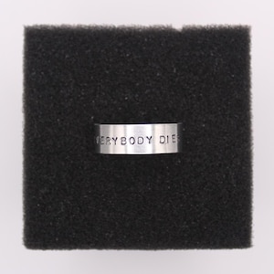 Könnte beinhalten: Ein silberner Ring mit der Aufschrift "EVERYBODY DIES" auf dem Band. Der Ring wird auf einer schwarzen, strukturierten Oberfläche präsentiert.