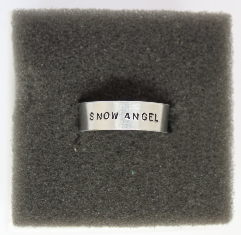 Puede incluir: Un anillo de plata con las palabras "SNOW ANGEL" grabadas.