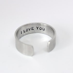 Könnte beinhalten: Ein offener Ring aus silberfarbenem Metall mit einer glatten, gebürsteten Oberfläche. Auf der Innenseite des Rings befindet sich der eingestanzte schwarze Schriftzug I LOVE YOU, der entlang des inneren Umfangs des offenen Rings sichtbar ist.