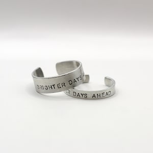 Può includere: Due anelli a fascia aperta in metallo color argento. Ogni anello presenta il testo nero stampato "BRIGHTER DAYS AHEAD" sulla superficie esterna. Il metallo ha una finitura spazzolata opaca e gli anelli sono posizionati in modo sovrapposto.