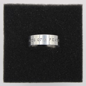 Könnte beinhalten: Ein silberner Ring mit dem eingravierten Schriftzug "BIRDS OF A FEATHER" auf der Außenseite. Der Ring ist vor einem schwarzen, strukturierten Hintergrund platziert. Der Ring hat ein schlichtes Banddesign.