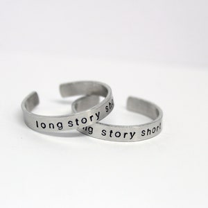 Könnte beinhalten: Zwei silberne Ringe mit der Gravur "long story short".