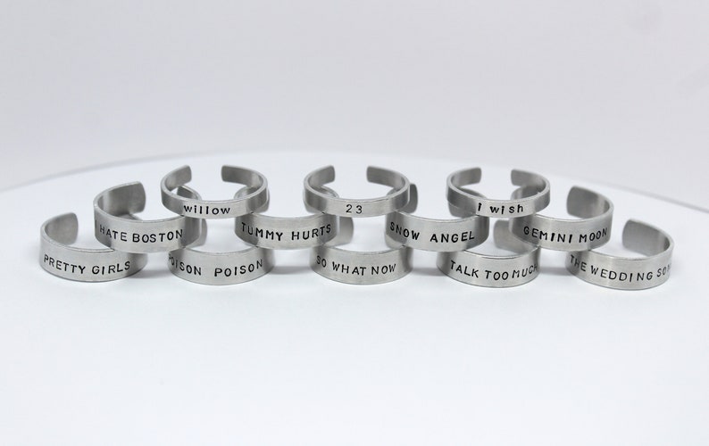 Puede incluir: Diez anillos de metal plateado con diferentes frases grabadas. Las frases incluyen "Pretty Girls", "Hate Boston", "Willow", "Digon Poison", "Tummy Hurts", "23", "So What Now", "Snow Angel", "I Wish", "Gemini Moon", "Talk Too Much" y "The Wedding So".
