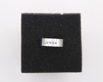 May include: A small, rectangular, silver-colored ring with the word "LUNCH" stamped on it, centered on a black, textured square. The background is plain white.