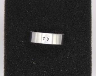 Puede incluir: Un anillo plateado con las letras "T 8" estampadas se muestra sobre un fondo negro. El anillo tiene un diseño de banda simple y plana. El anillo probablemente está hecho de metal.