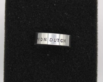 Puede incluir: Un anillo plateado con el texto "VON DUTCH" grabado en la banda. El anillo se muestra sobre una superficie negra, creando un contraste que resalta su diseño. El anillo es una pieza de joyería.