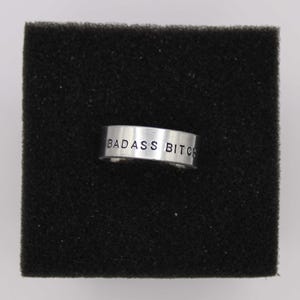 May include: A silver-colored ring with the words "BADASS BITCH" stamped on the outside. The ring is set against a black, textured background.