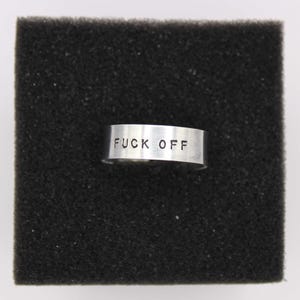 Peut inclure: Une bague argentée avec l'inscription "FUCK OFF" gravée sur le bandeau. La bague est présentée sur une surface noire et texturée.