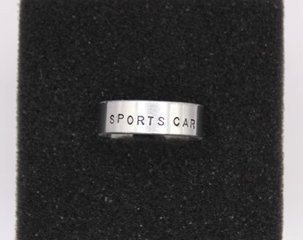 Puede incluir: Un anillo plateado con las palabras "SPORTS CAR" estampadas en la parte delantera. El anillo está sobre un fondo negro texturizado. El anillo es de estilo banda.