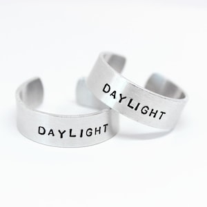 Könnte beinhalten: Zwei silberne Ringe mit der Gravur "DAYLIGHT" auf jedem. Die Ringe sind übereinander gestapelt.