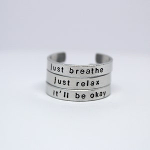 May include: A silver ring with three bands, each engraved with a different phrase: "just breathe", "just relax", and "it'll be okay".