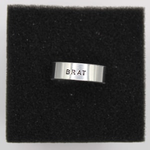 Könnte beinhalten: Ein silberner Ring mit dem Wort "BRAT" auf der Vorderseite. Der Ring ist vor einem schwarzen, strukturierten Hintergrund platziert. Der Ring scheint aus glänzendem Metall zu bestehen.